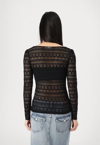Top nero in pizzo a maniche lunghe con motivi floreali e geometrici, caratterizzato da una silhouette aderente e un collo a barca. Indossato con jeans azzurri chiari.