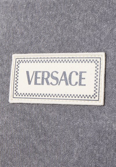 Fundo de tecido cinza com um rótulo retangular tecido, com "VERSACE" em letras maiúsculas em negrito, bordado por um padrão de quadrados branco e preto.