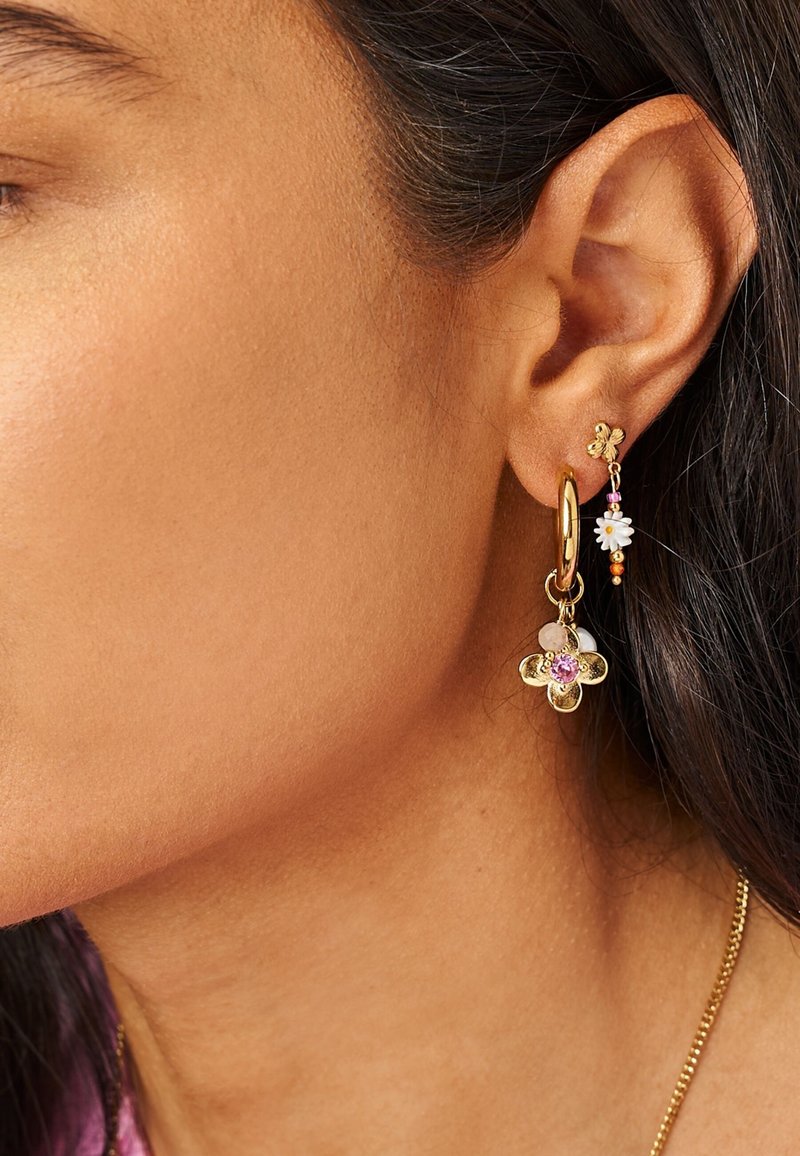 Primer plano de la oreja de una persona con dos pendientes de oro, uno con un colgante en forma de flor de gema rosa y el otro con un colgante de margarita blanca.