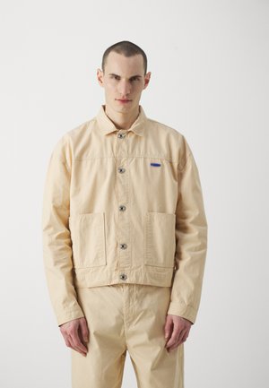 Veste beige clair en coton avec des fermetures à boutons, deux poches avant et une petite étiquette bleue sur la poitrine. Design simple, coupe décontractée.