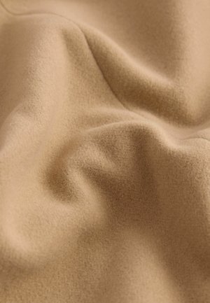 Weicher beiger Stoff mit leicht strukturierter Oberfläche, der sanfte Falten und Knicke zeigt. Das Material wirkt warm und gemütlich und eignet sich für Kleidung oder Polsterung.