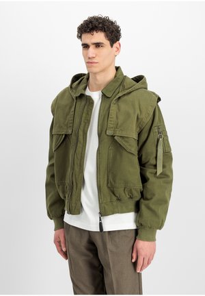 Jonge man met krullend haar die een olijfgroene bomberjas met capuchon draagt over een wit shirt en een bruine broek, staand tegen een witte achtergrond.