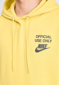 Sárga kapucnis pulóver, zsinórokkal, amelyen a "HIVATALOS HASZNÁLATRA" felirat és a Nike logó sötétszürke színben látható. Pamut anyag, sima textúrával.