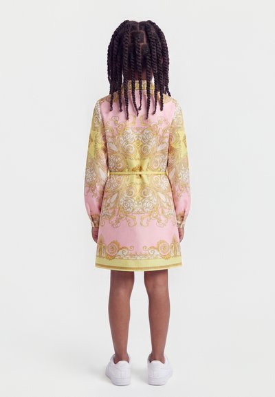 Κορίτσι με πλεξούδες στέκεται πλάτη, φοράει ροζ και κίτρινο φόρεμα με διακοσμητικό σχέδιο και λευκά αθλητικά παπούτσια σε απλό φόντο.