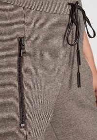 Pantalon à carreaux marron et beige avec taille à cordon, poche zippée à l'avant et tissu texturé. Présente des accents de quincaillerie noire.
