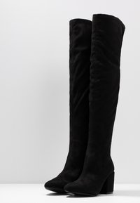Stivali alti neri in suede con tacco a blocco e punta affusolata, in posizione verticale su una superficie bianca uniforme contro uno sfondo grigio chiaro.