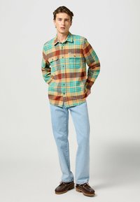 Camisa de cuadros con tonos de marrón, azul verdoso y amarillo. Mangas largas, dos bolsillos frontales y un diseño abotonado, combinado con jeans azul claro.