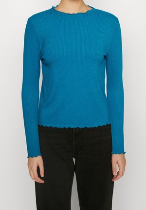 Gestreiftes Langarmoberteil in leuchtendem Blau mit einem gewellten Ausschnitt und Saum, aus einem strukturierten Material gefertigt. Getragen mit dunklen Hosen.