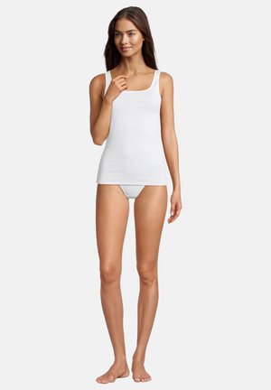 Junge Frau steht barfuß, trägt ein weißes Tanktop und weiße Unterwäsche vor einem schlichten Hintergrund.