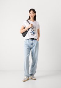 T-shirt blanche avec un imprimé graphique représentant une ville ; associée à un jean en denim bleu clair et des baskets argentées ; sac à dos noir.