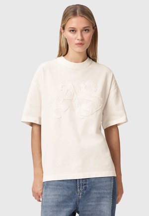 Ung kvinde med glat blondt hår iført en cremefarvet oversized T-shirt med teksturerede "AS"-bogstaver og blå jeans, stående mod en ensfarvet baggrund.