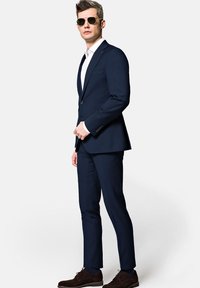 Granatowy garnitur o dopasowanym kroju, z klapami typu notch, zapięciem na jeden guzik i gładkim materiałem. Połączony z białą koszulą i ciemno-brązowymi butami.