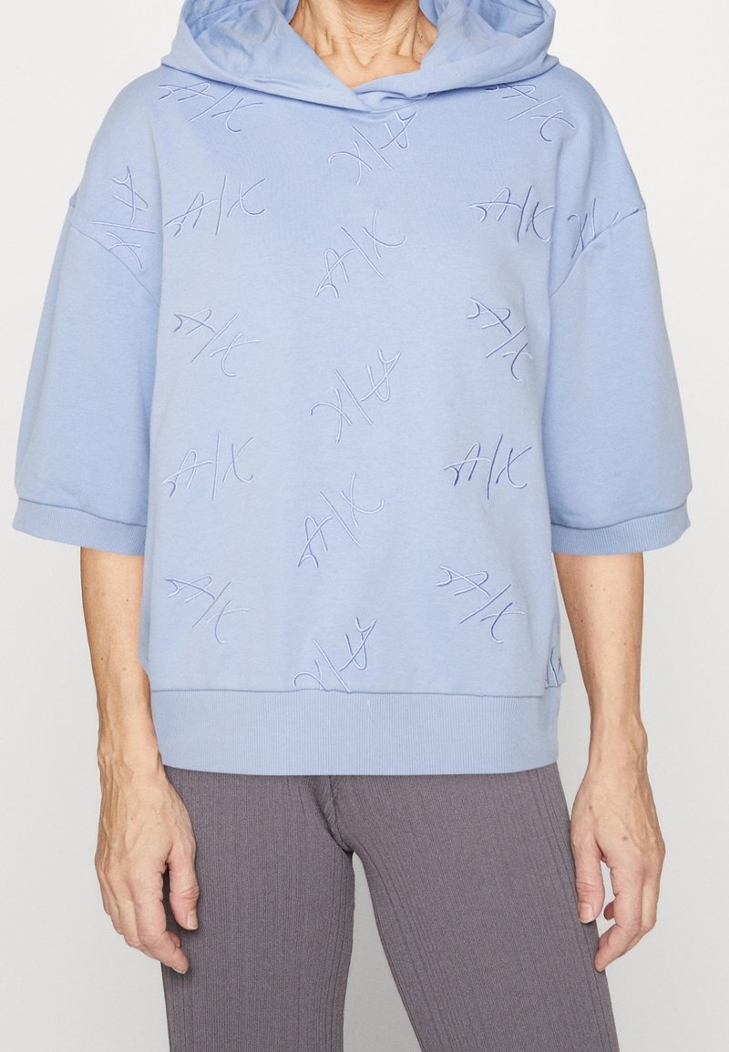 Osoba oblečená v světle modré mikině s kapucí a krátkými rukávy, na které se opakovaně nachází vyšívané logo "A|X", doplněná o žebrované šedé kalhoty.