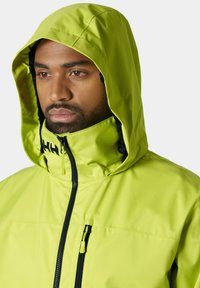 Chaqueta impermeable verde brillante con capucha ajustable, cremallera negra y bolsillo en el pecho. Textura suave con un diseño elegante y funcional.