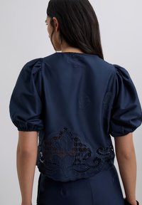 Blusa de manga abullonada azul marino con intrincados detalles de encaje en el dobladillo, que presenta bordados florales y una textura suave similar a la satén.