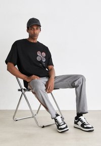 T-shirt nera con grafica, caratterizzata da stampe circolari multicolori, abbinata a pantaloni grigi con strisce laterali arancioni e sneaker in bianco e nero.