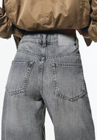 Jeans din denim gri cu talie înaltă, două buzunare pe spate și un eticheta de marcă vizibilă pe talie. Materialul are o textură decolorată.