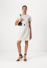 Robe à manches courtes de couleur beige clair avec un motif géométrique, associée à un sac à main noir et des talons noirs à lanières. Équipée d'une ceinture à la taille.