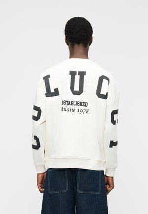 Personne portant un sweat-shirt blanc avec les inscriptions "LUC" et "Established Milano 1978" en noir, associé à un jean large bleu foncé.