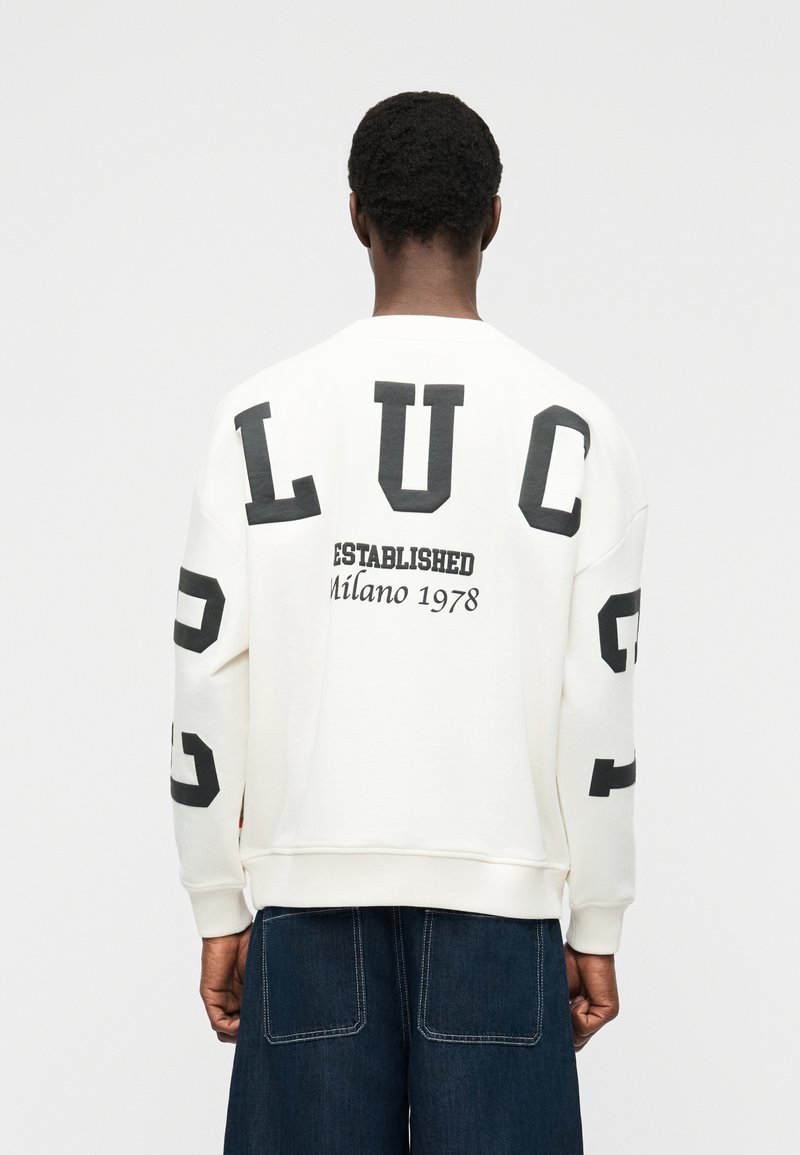 Personne portant un sweat-shirt blanc avec les inscriptions "LUC" et "Established Milano 1978" en noir, associé à un jean large bleu foncé.
