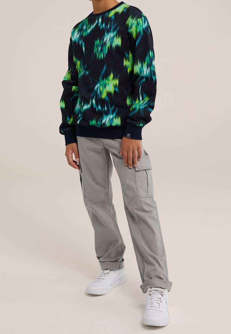 Personne portant un sweat-shirt noir à manches longues avec des motifs abstraits bleus et verts, un pantalon cargo gris clair et des baskets blanches.