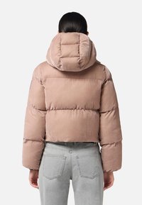 Chaqueta acolchada corta de tejido rosa suave, con una gran capucha y detalles de costura, combinada con pantalones vaqueros de talle alto en gris claro.