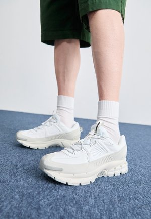 Jambes en shorts verts et chaussettes blanches hautes, portant des baskets blanches épaisses, debout sur un sol recouvert de moquette bleue, contre un mur blanc uni.