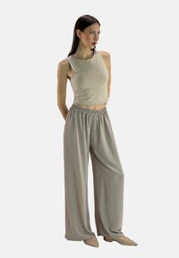 Beige mouwloos topje met een getailleerd ontwerp, gecombineerd met losse, lichtgrijze broek met elastische taille. Model draagt nude-kleurige platte schoenen.