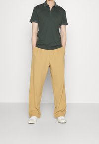 Polo vert foncé à col zippé, associé à un pantalon plissé beige clair et des baskets blanches. Tissu lisse avec un détail de col contrastant.