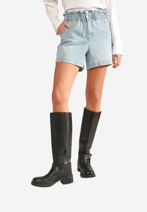 Frau trägt hellblaue Denim-Shorts mit Paperbag-Taille, Vorderknöpfen und schwarze kniehohe Lederstiefel, steht mit einer Hand in der Tasche.