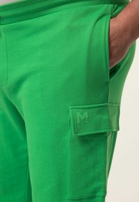 Groene sweatpants gemaakt van zachte stof, met een zijvak van het type cargopocket met een reliëflogo. Glad textuur en een elastische tailleband.