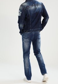 Man i en sliten blå denimjacka och jeans, stående med händerna i fickorna, vänd bort från kameran mot en neutral bakgrund.