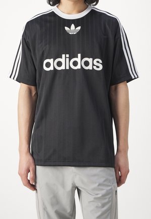 Person iført en sort Adidas t-shirt med hvidt logo og striber på ærmerne, kombineret med lysegrå bukser.