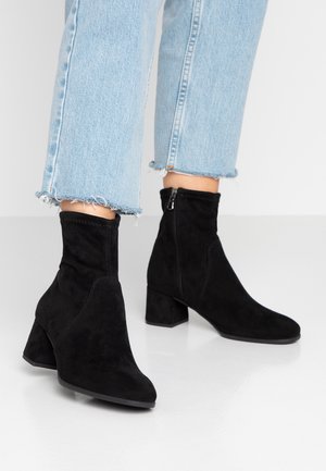 Bottes de cheville en suede noir avec talons carrés portées avec un jean bleu clair à ourlet effiloché sur un sol blanc.