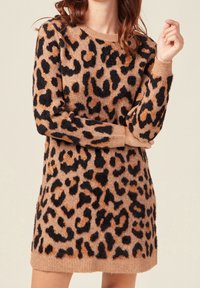 Femme portant une robe-sweater à manches longues, arrivant aux genoux, avec un motif léopard brun, noir et orange.