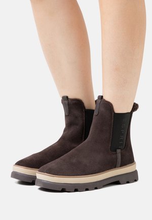 Botas de tobillo de ante marrón oscuro con suela beige y oscura, paneles laterales elásticos, usadas sobre piernas desnudas contra un fondo blanco.