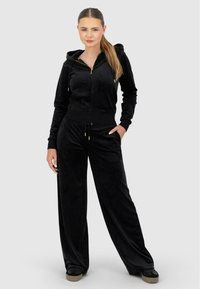 Melna velūra treniņtērps ar džemperi ar rāvējslēdzēju, priekšējām kabatām un regulējamu jostu, papildināts ar zelta aksesuāriem.