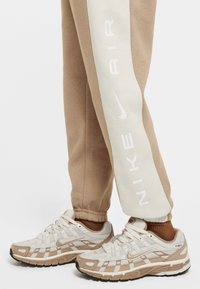 Běžecké boty Nike v béžové a krémové barvě s vrchní částí ze síťoviny, gumovou podrážkou a reflexními akcenty. Béžové joggers kalhoty s bílým postranním panelem a logem.