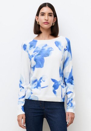 Kobieta o prostych ciemnych włosach, nosząca złote kolczyki koła i biały sweter w niebieskie kwiaty, do tego ciemnoniebieskie jeansy.