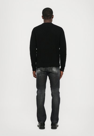 Pull en tricot noir avec une texture côtelée, légères déchirures à l'ourlet, associé à un jean gris foncé et des chaussures noires, vu de dos.