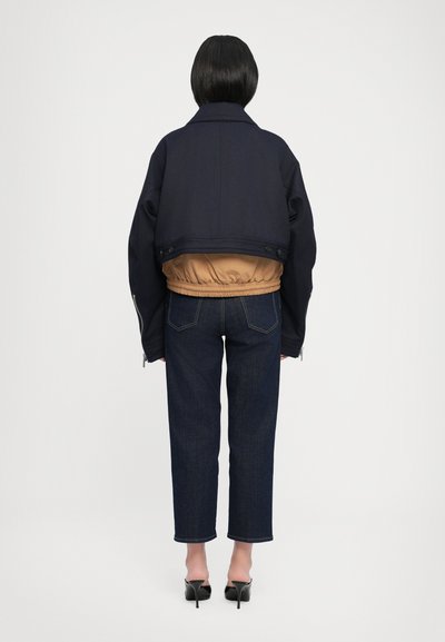 Chaqueta navy cortada con un forro interior beige, combinada con pantalones de mezclilla oscuros de pierna recta. El modelo lleva zapatos de tacón negros, visto desde atrás.