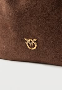 Brązowa zamszowa torebka z zapięciem w kolorze złotym w kształcie dwóch ptaków z rozpostartymi skrzydłami. Gładka faktura i minimalistyczny design. Widoczne logo marki.