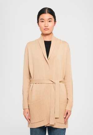 Jonge vrouw in een beige gebreide cardigan met sjaalkraag, voorzakken en een strikceintuur over een zwart topje en blauwe jeans.