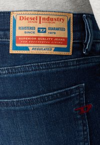 Detalhe de uns jeans de ganga azuis com um rótulo de couro castanho a dizer "Diesel Industry Denim Division" e um pequeno logótipo bordado em vermelho no bolso.
