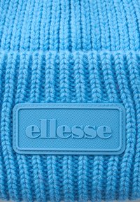 Mėlyna megzta medžiaga su mėlynu guminio ženklelio emblema, iškilminiuoju būdu užrašyta prekės ženklo pavadinimu "ellesse."