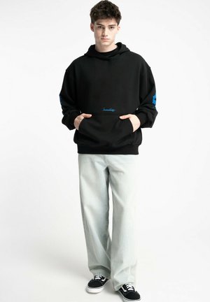 Czarna bluza z kapturem z kieszenią z przodu, niebieskie akcenty na rękawach, w zestawie z jasnymi dżinsami i czarnymi butami do skate'a. Casualowy fason i luźny krój.