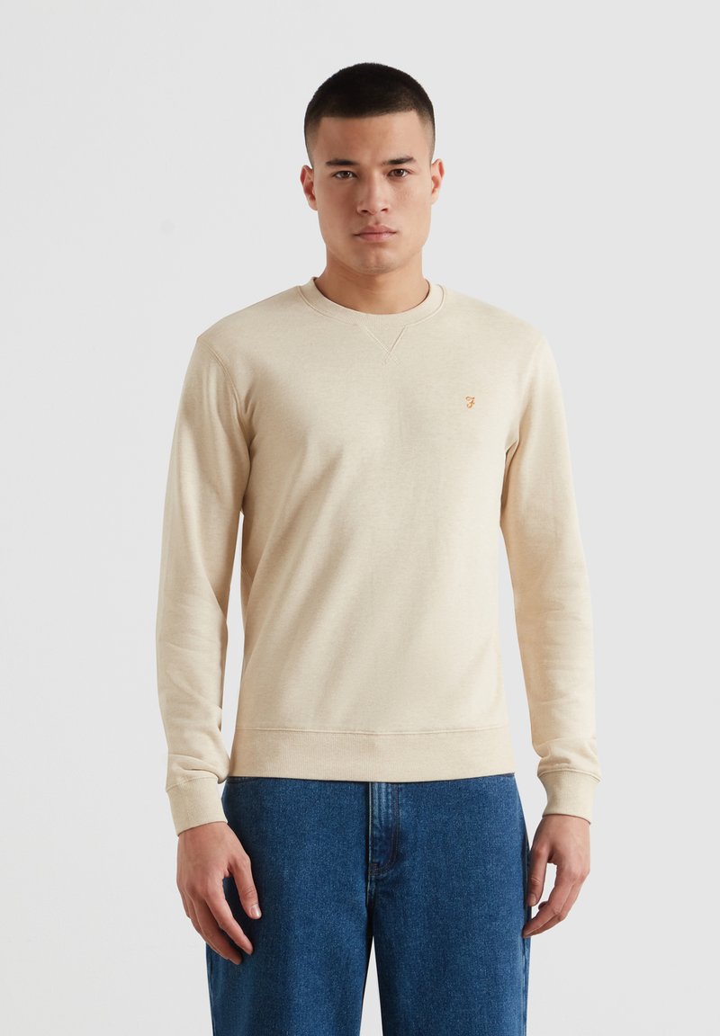Homme portant un sweat-shirt crème à manches longues et un jean bleu, debout devant un fond uni gris clair, faisant face à l'appareil avec une expression neutre.
