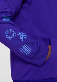 Sudadera morada con diseños gráficos azules en las mangas, que incluyen símbolos y patrones. El material es suave y tiene una textura lisa.