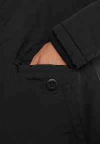 Mustassa takissa on näkyvä tasku. Kankaan pinta on teksturoitu. Tasku suljetaan mustalla napilla, jossa on kohokuvioituja yksityiskohtia.