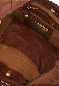 Interno di borsa in pelle marrone con una trama liscia, fodera color cuoio, tasca con cerniera e targhetta con marchio. Accenti in ottone sugli angoli.